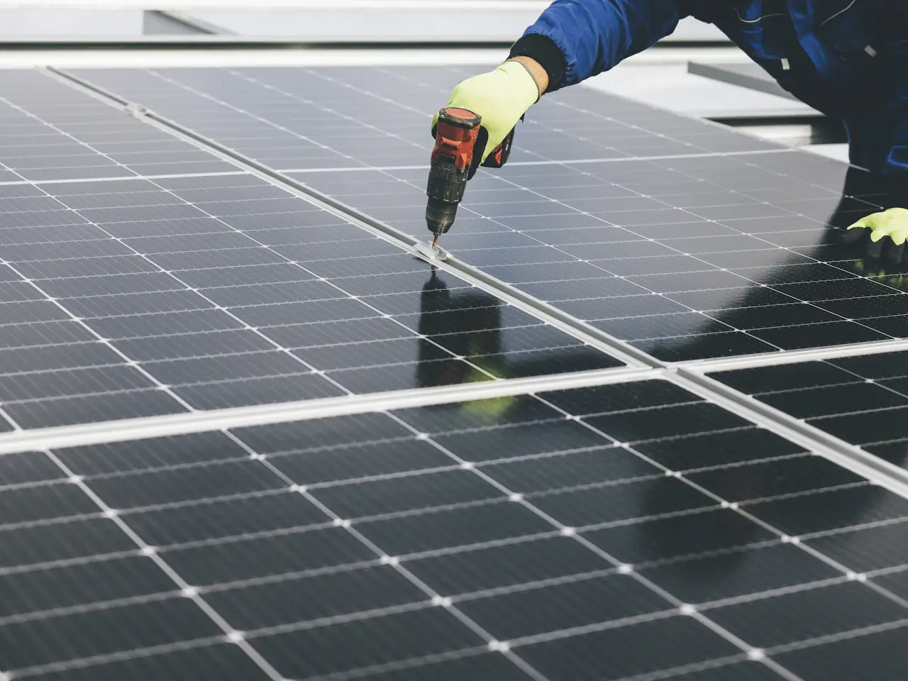 Monteur in blauer Arbeitsjacke und Schutzhandschuhen zieht mit einem Akkuschrauber eine Modulklemme zwischen zwei Photovoltaikmodulen auf einem Dach fest