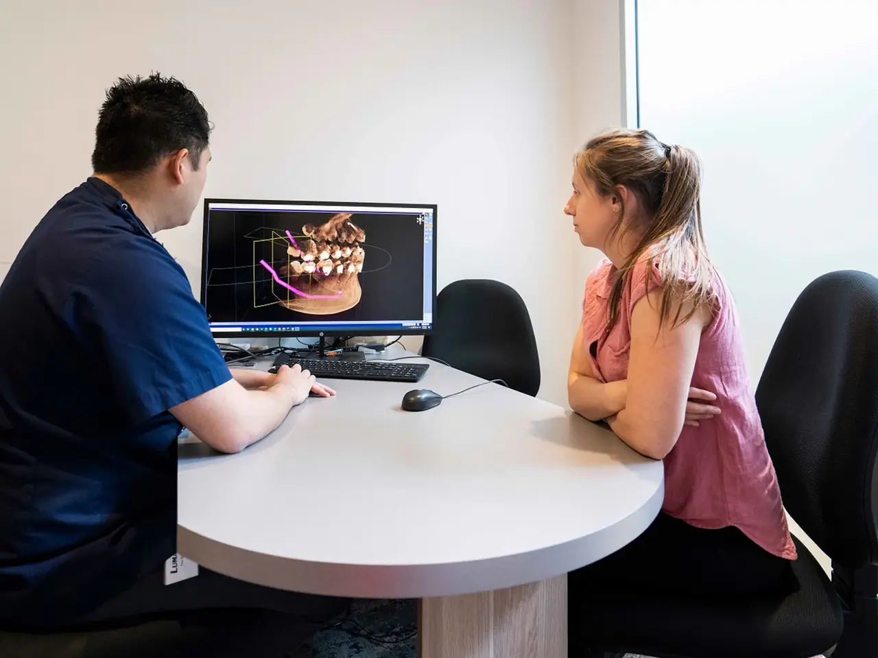 Zahnarzt und Patientin sitzen an einem runden Tisch und besprechen am Bildschirm eine 3D-Aufnahme des Kiefers für die Behandlungsplanung