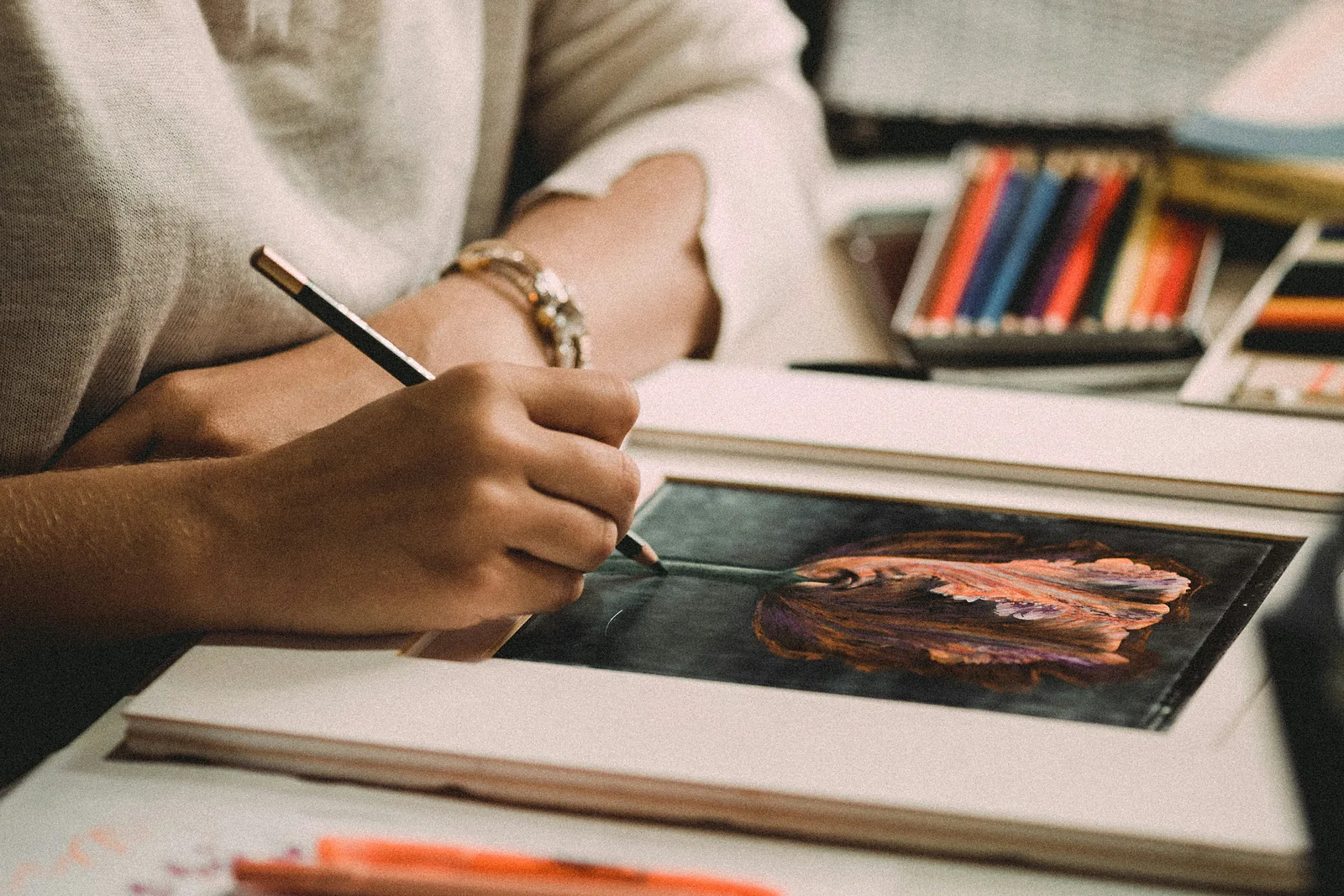 Hand zeichnet mit einem Bleistift eine Tulpe auf dunklen Karton, dahinter ein offenes Etui mit Farbstiften