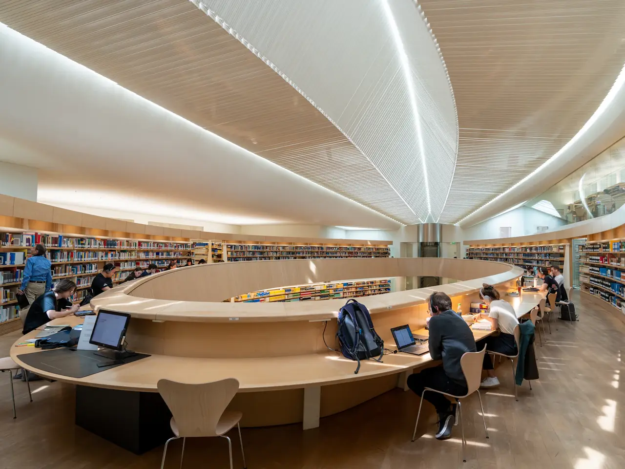 Studierende arbeiten an Laptops und mit Büchern an einem geschwungenen Holztisch in einer hellen Bibliothek mit langen Bücherregalen