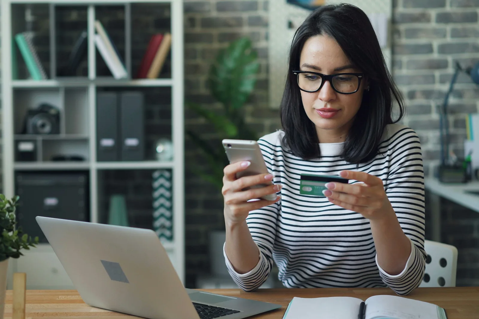 Eine Frau mit Brille bestellt am Schreibtisch mit Smartphone und Kreditkarte, vor ihr ein aufgeklappter Laptop