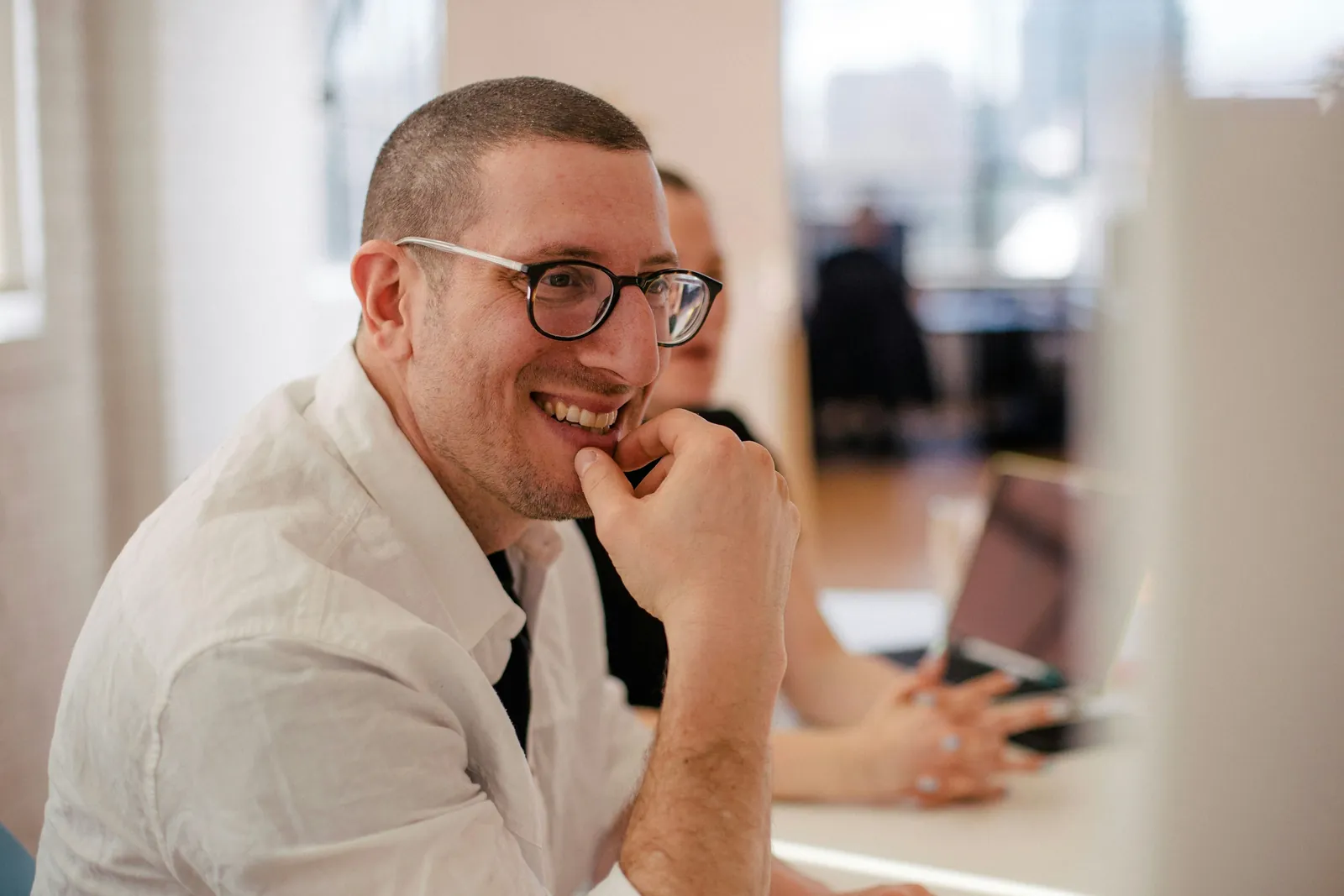Ein Mann mit Brille lacht im Gespräch an einem Schreibtisch in einem hellen Büro