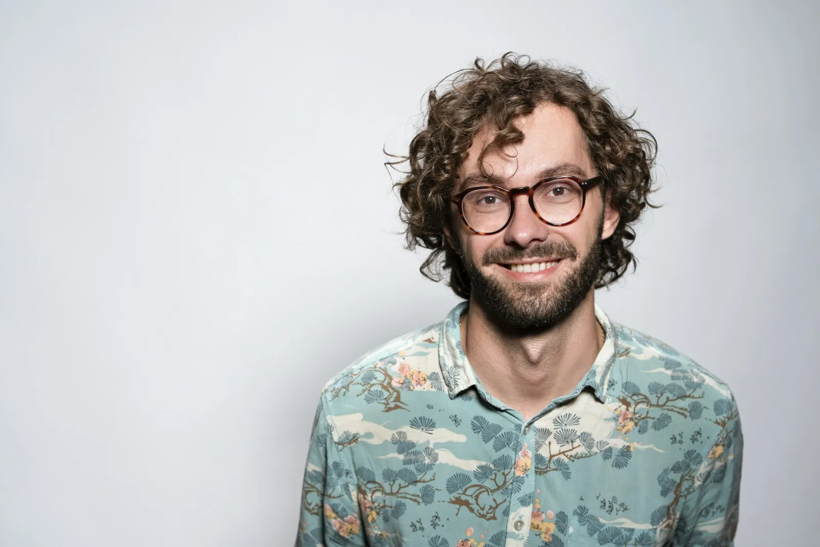 Porträt eines lächelnden Mannes mit Brille und Locken vor grauem Hintergrund