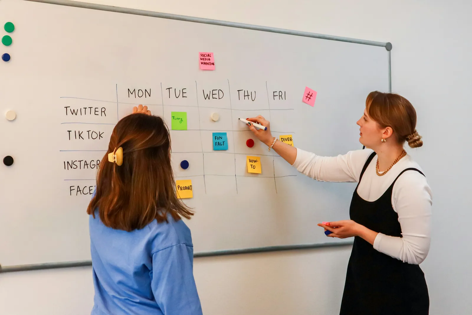 Zwei Frauen planen am Whiteboard Social-Media-Beiträge für die Woche, eine schreibt, die andere schaut zu