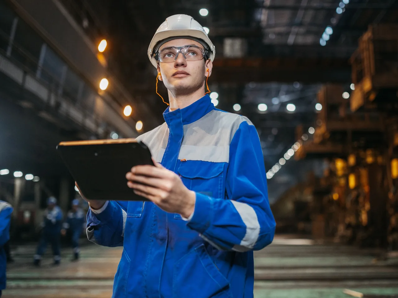 Techniker mit Helm und Schutzbrille hält in einer Werkhalle ein Tablet in der Hand