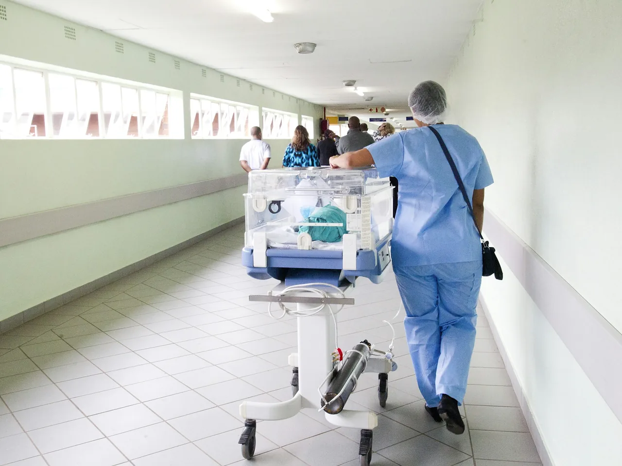 Pflegekraft in blauer Klinikkleidung schiebt einen Inkubator durch einen hellen Krankenhausflur